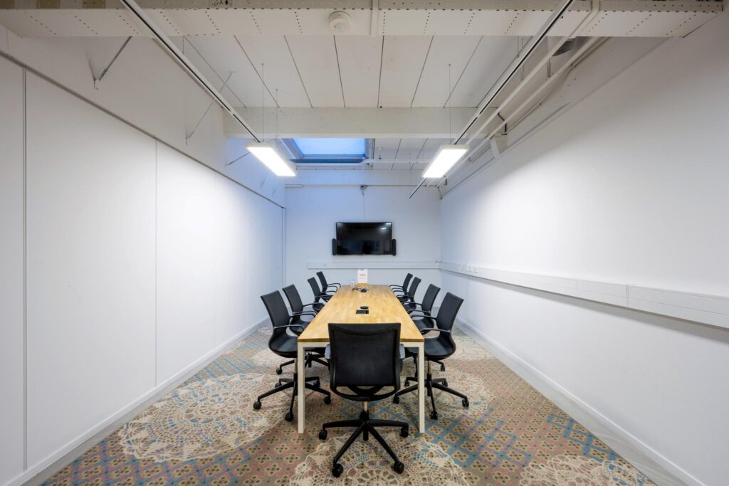 Vergaderruimte aan de Van der Madeweg met een lange tafel, tien stoelen, televisie en kleurrijk tapijt.